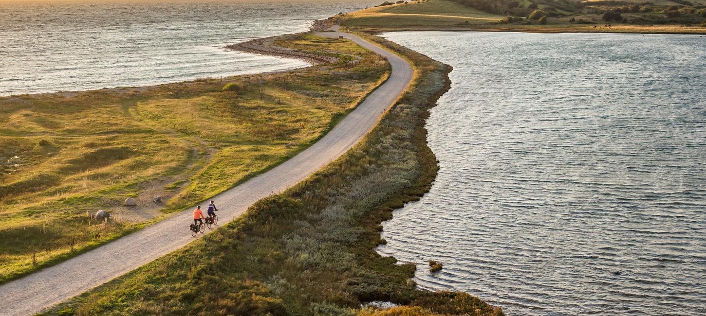 Luftfoto af to cyklister på smal vej ud af tange med vand på begge sider og land i baggrunden.