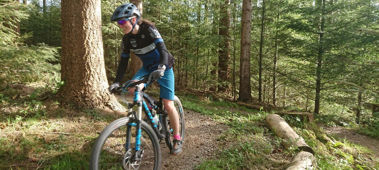 En kvinde i blåt cykeltøj cykler på et mountainbike-spor. Hun er på vej rundt i et sving og er omringet af skov.