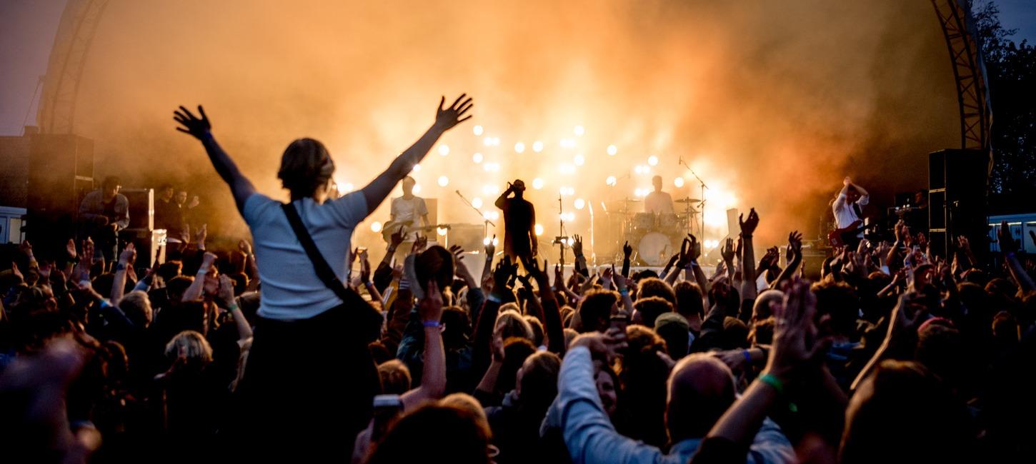 Foran en scene står et band oplyst af orange lys. Publikum står med begge hænder i vejret. En kvinde sidder på skulderen af en anden og hun har begge arme over hovedet. 