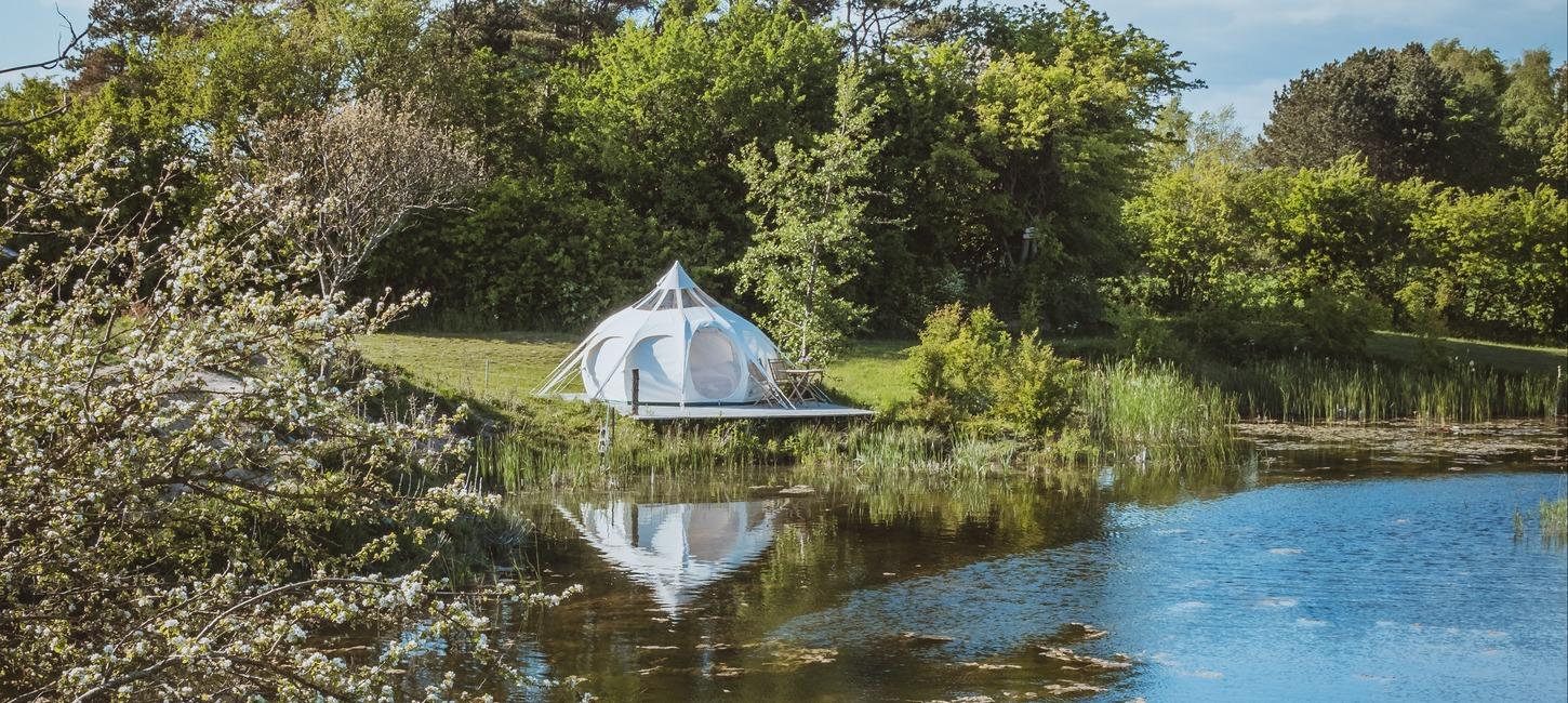 Hvidt glampingtelt i naturskønne omgivelser ned til vandet.