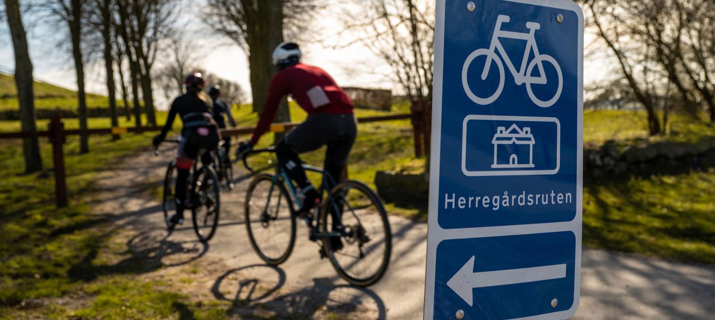 i forgrunden er et blåt skilt med en cykel, pil til venstre og tekst hvor der står Herregårdsruten. I baggrunden tre personer på racercykel ned af en sti der går mellem træer. 