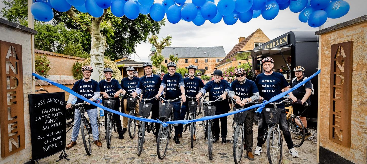 De fynske borgmestre stå klar på cyklen på ræd og række foran et blår bånd ved Andelen på Ærø. En port af blå balloner ses henover dem.