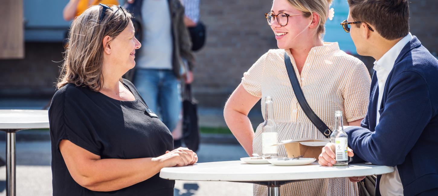 Tre mennesker, der netværker ved et højt cafebord udenfor i solskinnet.