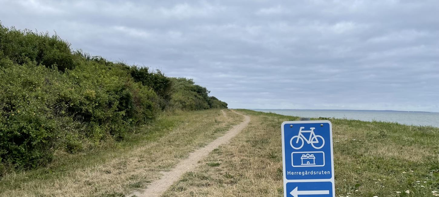 En græseng der grænser op til havet med en lang grussti som fortsætter i horisonten. I forgrunden er et blåt skilt med en cykel, en pil til venstre og navnet Herregårdsruten. 