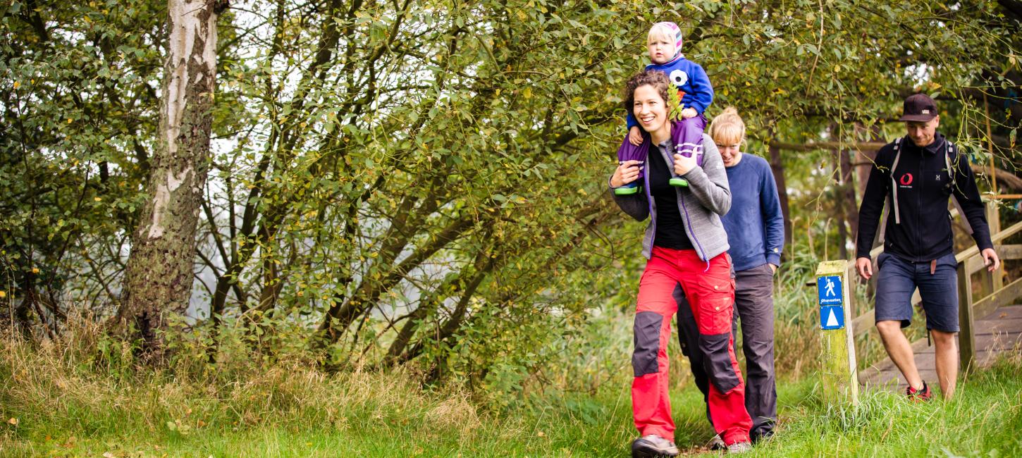 Familie på vandretur i en skov. Forrest er en person med et barn på skuldrene og bagved ses to yderligere personer samt et blåt vandreskilt.