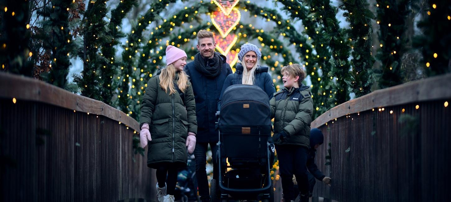 På en træbro står en familie med tre brøn i vintertøj. Foran moren står der en barnevogn. Over hele broen er der buer med gran, lyskæder og i midten et lysende julehjerte.