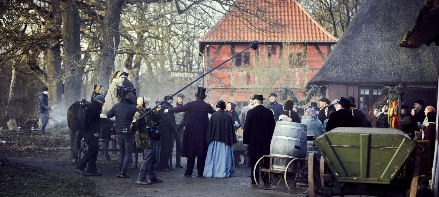 Et billede fra en filmoptagelse hvor der står både en mand med mikrofonstang og en mand med et kamera. De filmer en masse mennesker på en grusvej som står i gammeldags tøj. De står mellem to bindingsværkshuse. Til højre på billedet står en grøn hestekærre.