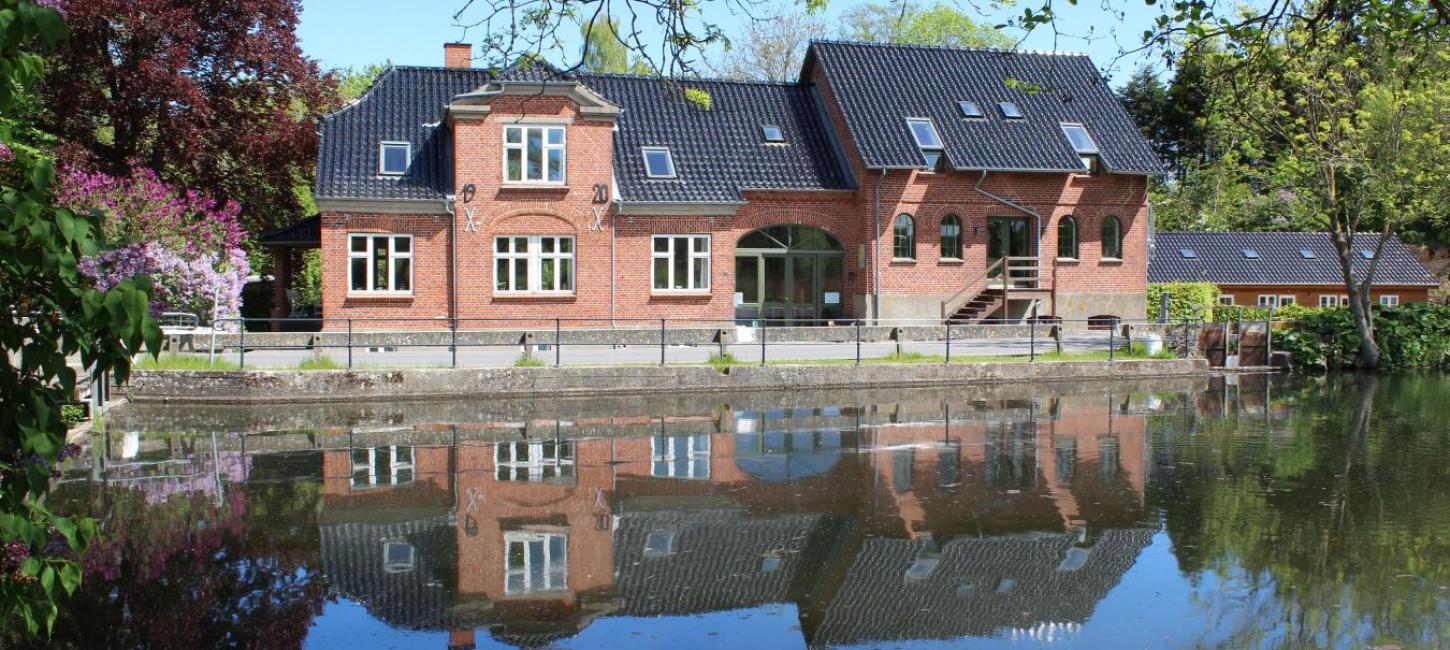 Stort, rødt hus ligger ud til noget vand og spejler sig flot i det. 