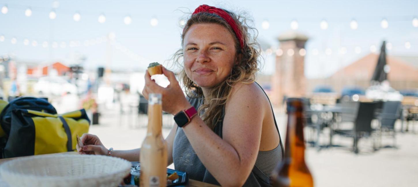 Kvinde med snack i hånden sidder med og mad og drikke på bordet foran og smiler til kameraet.