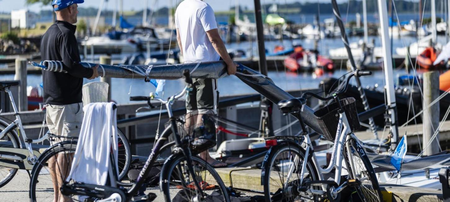 To cykler står parkeret på Middelfart Marina. I baggrunden er to sejlere i gang med at bære et sammenrullet sejl ned på en sejlbåd.