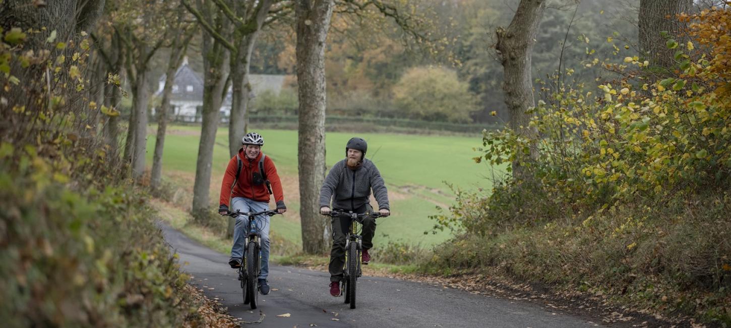 To cyklister cykler på asfaltvej omgivet af marker og træer i efterårets farver.