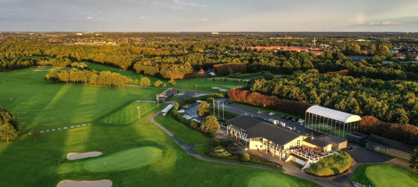 Udsigt over grøn golfbane oppefra med golfklubbens lokaler i forgrunden og Odense by i baggerunden.