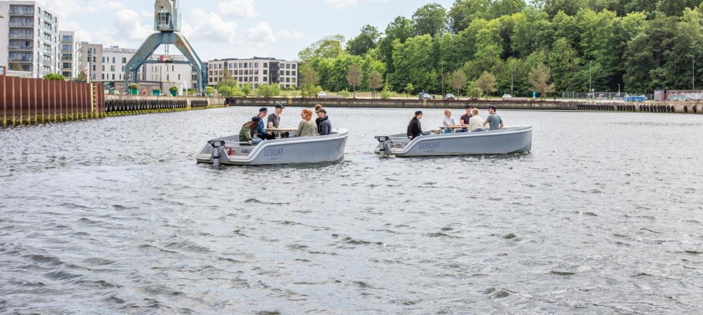 To GoBoat-både på vandet ved Odense Havn med grønne træer og en stor kran i baggrunden