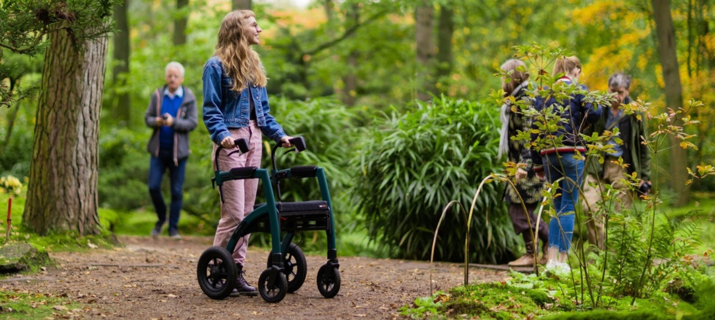 Ung pige med rollator står i grønne omgivelser med andre mennesker i baggrunden