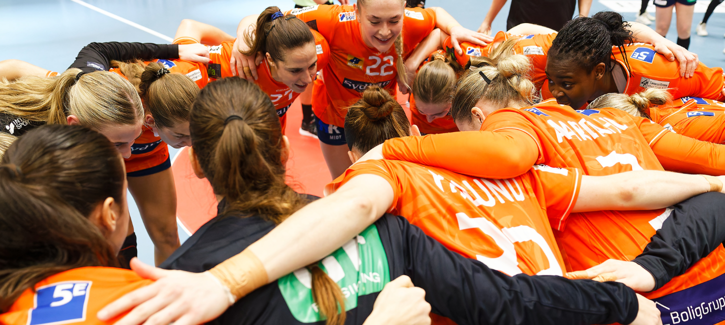 Håndboldkvinder i orange spillertøj danner rundkreds med armene om hinanden