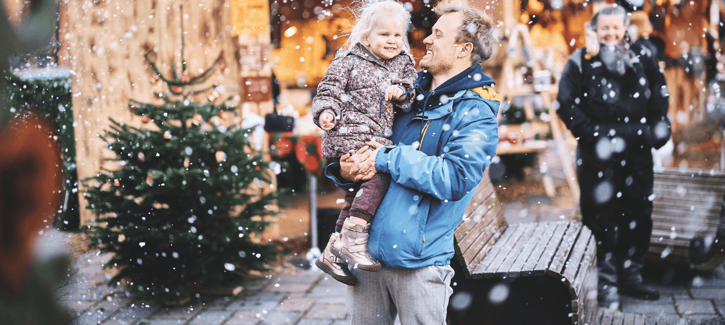 En mand holder et smilende barn i sine arme, mens han står udenfor i sneen, tæt på et juletræ og festlige julemarkedsboder.