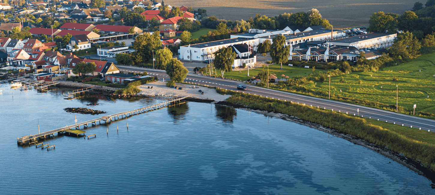 Dronefoto af hotel lige ud til kysten med grønt græs, blåt hav og et rødt svajende dannebrog
