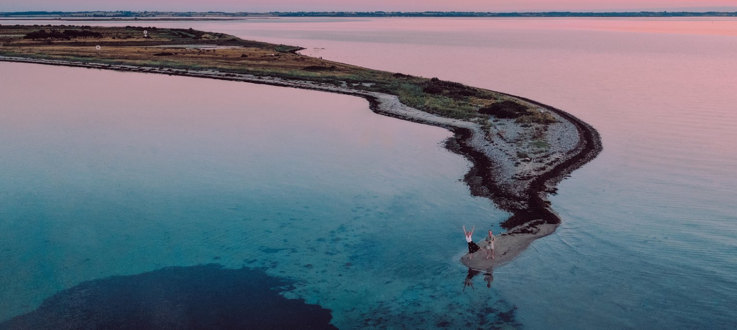 Et par står for enden af en landtange på et dronebillede med solnedgangen i baggrunden