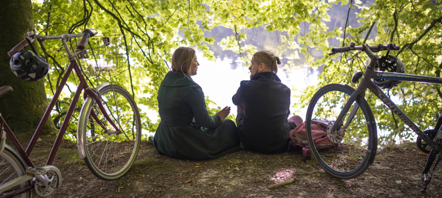 Par sidder i skovbrynet med ryggen til. En cykel er parkeret på hver side af dem.