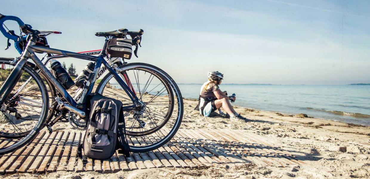 To parkerede cykler på strand. I baggrunden sidder de to cykelryttere i sandet i vandkanten og holder pause.