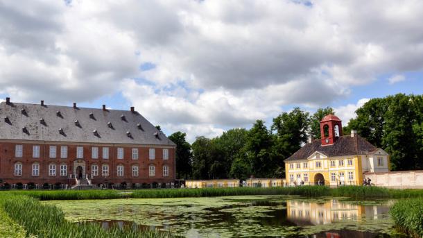 Valdemars Slot hovedbygning og porthus på en sommerdag med overskyet himmel. I forgrunden ses gårdspladsens vandbassin.