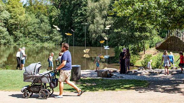 Mand med barnevogn går forbi børn, der leger ved vand med store kunstsommerfugle på pinde.