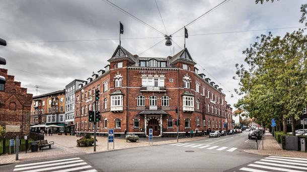 Stor, rød, symmetrisk, slotsagtig bygning i bymiljø med fodgængerfelter og lyssignaler i forgrunden under en mørk og faretruende himmel.