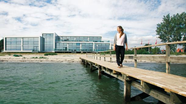 En dame går langs en badebro og kigger ud over vandet. Et stort hotel med vinduer på hele facaden ligger i baggrunden.