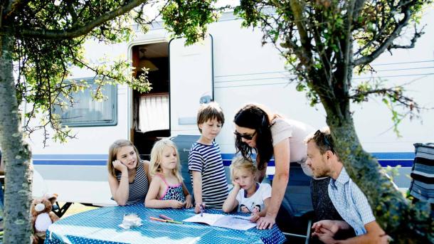 To voksne og fire børn samlet ved et campingbord foran en campingvogn