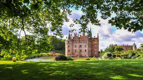 Egeskov slot med grønt græs i forgrunden og grønne træer og blå himmel i baggrunden.