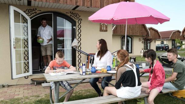 Ved et borde-bænke sæt sidder en familie og spiser aftensmad under en pink parasol. Ud fra sommerhuset i baggrunden kommer en mand med en skål i hånden. 