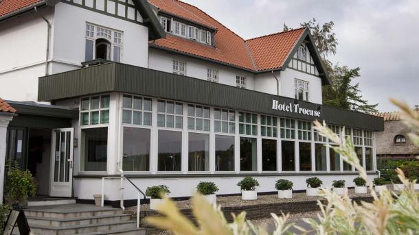 Hvid bygning med rødt tag. Ud fra bygningen er en hvid udestue med terrasse ovenpå. På terrassens sorte hegn står der Hotel Troense.