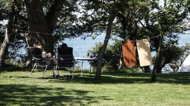 To mennesker sidder på en græsbakke på klapstol og kigger ud på havet. I baggrunden hænger to håndklæder til tørre på en snor og et hvidt telt er slået op.