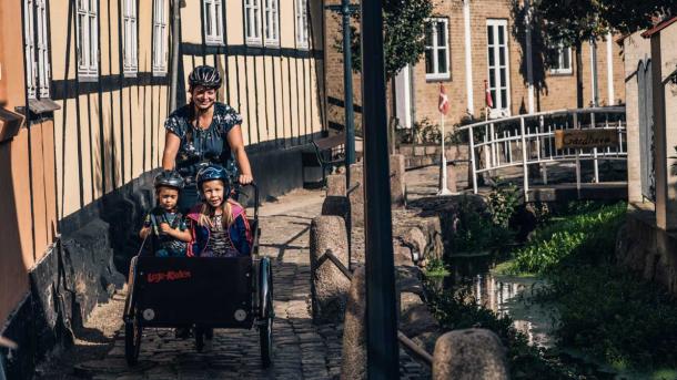 Kvinde kommer kørende på ladcykel med to børn i på en smal, brostensbelagt gade med Bybækken til den ene side og gamle bindingsværkshuse til den anden side.