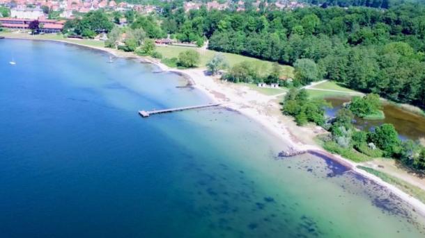 Strand set ovenfra med lyst sand og blåt og turkis vand.