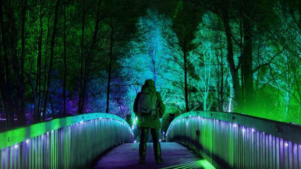 Midt på en bro står en mand i vintertøj. På hver side af ham er gelænderet på broen oplyst af blå og grønne lamper. I baggrunden ses halvoplyste træer.