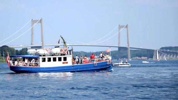 På havet foran Lillebæltsbroen sejler et blåt træskib med hvidt førerhus. På dækket står en flok mennesker og kigger udover vandet, og i bagenden flagre det danske flag.