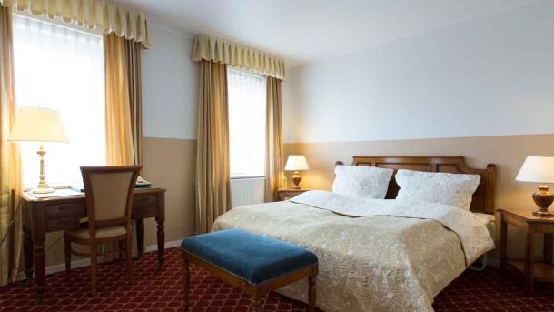 Et hotelværelse med en redt dobbeltseng, tændte lamper på bordene ved siden af sengen og et skrivebord foran et af vinduerne.