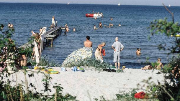 Strand med bølgende vand, badebro og mange badende gæster.