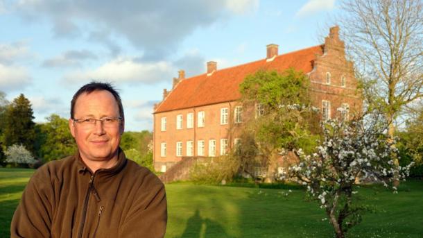 Mand med briller fotograferet foran et stort, rødt gods i baggrunden. 