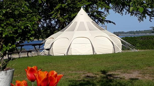 Stort hvidt glampingtelt på græsplæne i grønne omgivelser med blåt hav og blå himmel i baggerunden.