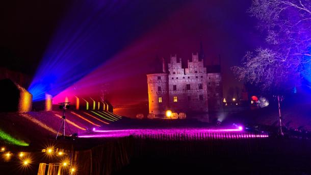 I baggrunden står Egeskov slot i mørket. Den lange græsplæne foran slottet er lyst op af forskellig farvede lamper. 