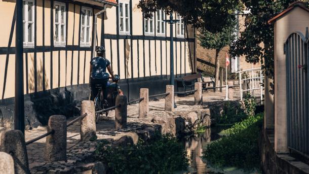 Cyklist trækker af sted med sin cykel på brostensbelagt sti langs med gult bindingsværkshus.