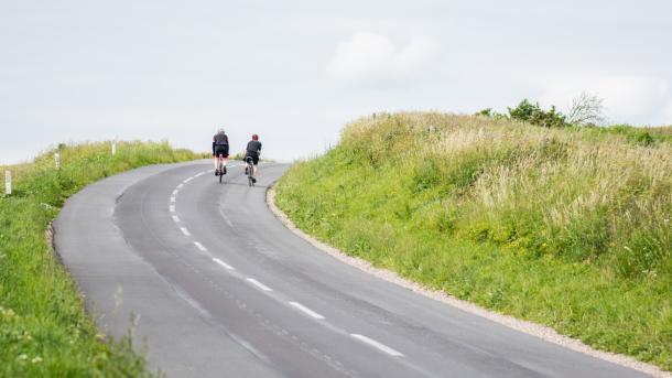 To cyklister på vej op ad bakke på landevej med grønt omkring. 