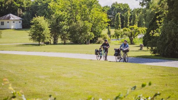 En par med hver deres cykel med opbakning går på en grussti. Foran stien er der græsplæne mens der bagved er græs og træer. 