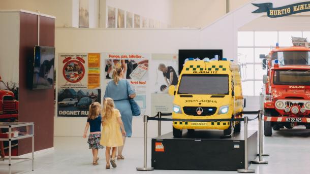 Ambulance og brandbil på indendørs udstilling. En mor med to børn går rundt og kigger på udstillingen.