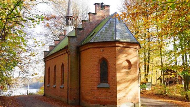 I en lysning mellem træer står et kapel i røde mursten. Træerne har alle efterårets farver og på asfalten rundt om kapellet ligger brune blade.