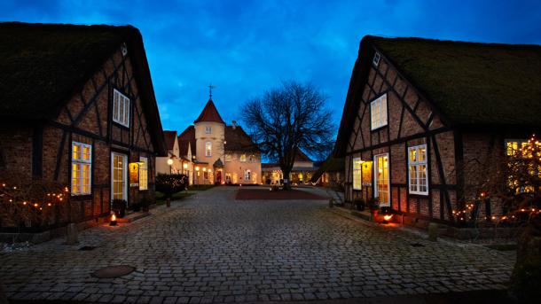 Stort bygningskompleks med stråtage, bindingsværk og julebelysning om aftenen.