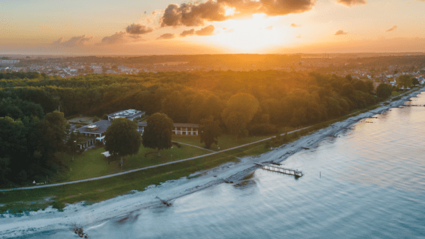 Et dronefoto af Hotel Hesselet, som ligger lige ud til kysten, set under en solnedgang med orange nuancer i horisonten bagved hotellet
