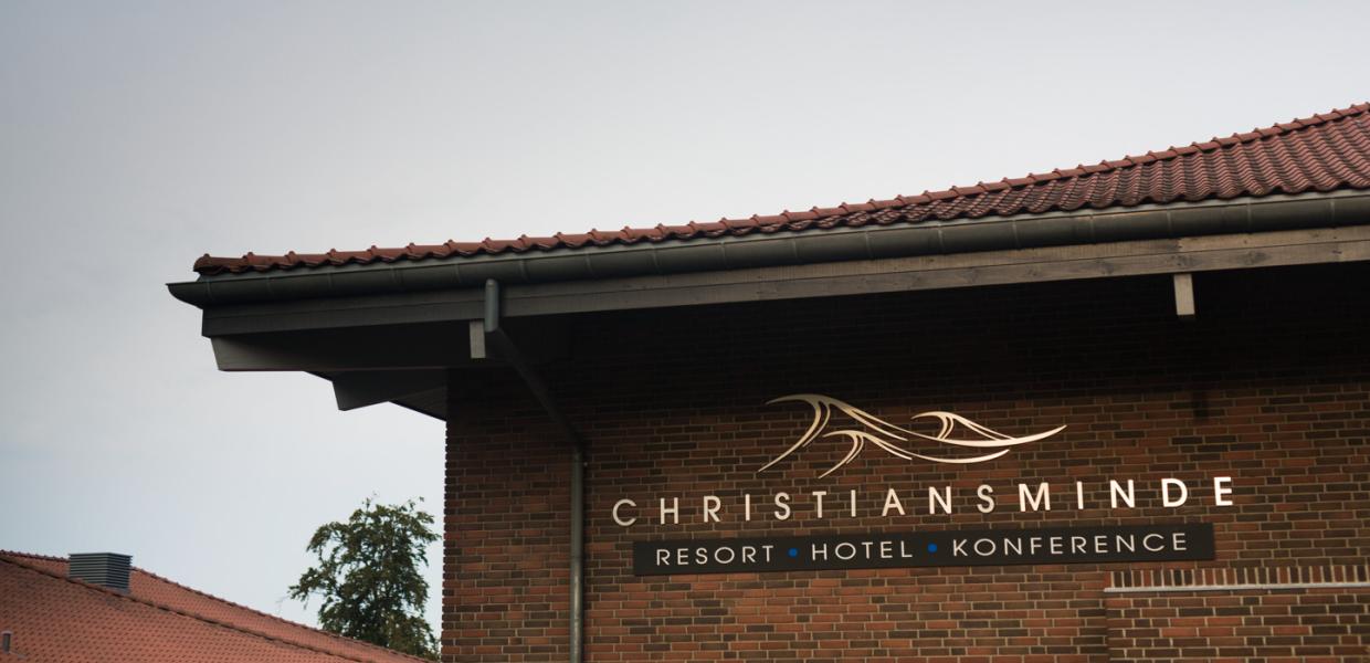 Et nærbillede af et hushjørne hvor logoet til Hotel og konference Christiansminde er i guld. Logoet er tre bølger hvorunder der står Christiansminde, resort, hotel, konference. 